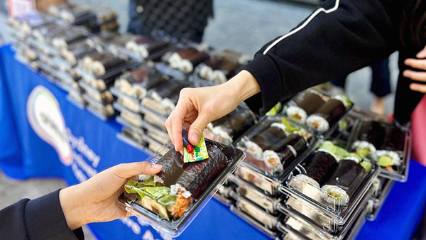 Photo of a hand passing out 2 sushi rolls in plastic packaging in front of a table with many sushi rolls